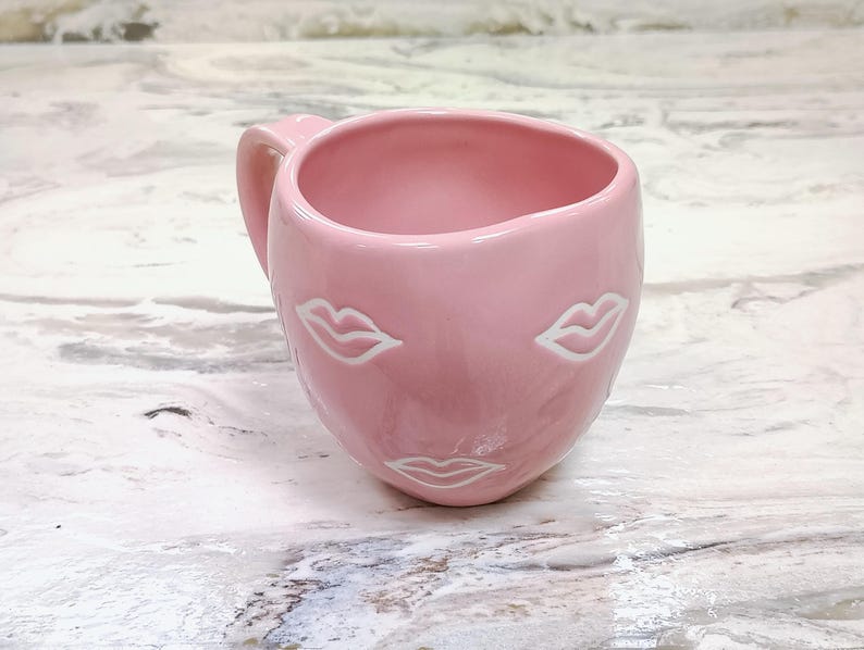 May include: A pink ceramic mug with a handle, featuring a face design with white lips. The mug has a smooth, glossy finish and is set against a marble-like surface. The mug is designed for beverages.