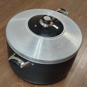 May include: A black cooking pot with a silver lid and a glass top. The pot has two handles and a knob on the lid. The pot is sitting on a wooden surface.