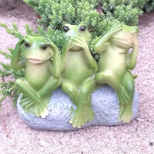 May include: A decorative figurine featuring three green frogs in a "see no evil, hear no evil, speak no evil" pose. The frogs are sitting on a gray stone base, with a natural background of greenery.