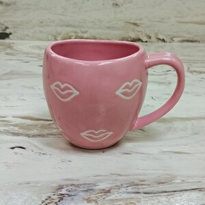 May include: A pink ceramic mug with a rounded body and a handle. The mug features three white lip designs. The mug is sitting on a light-colored surface.