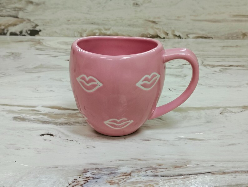 May include: A pink ceramic mug with a rounded body and a handle. The mug features three white lip designs. The mug is sitting on a light-colored surface.