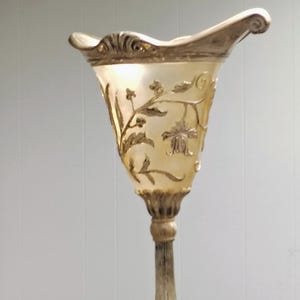 May include: A decorative, ornate vase with a flared top and a pedestal base. The vase is primarily a pale yellow color with gold-colored accents and floral designs. The base and rim have a metallic finish.
