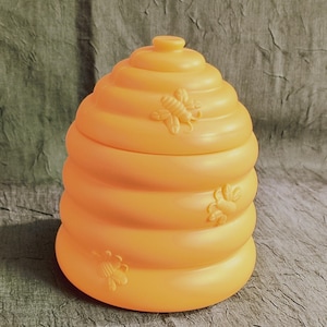 May include: A bright orange, beehive-shaped container with a lid. The container has a spiral design and is decorated with three embossed bee figures. The beehive is made of plastic and is sitting on a textured surface.