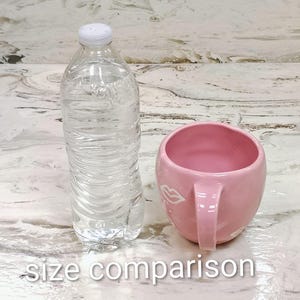 May include: A clear plastic water bottle with a white cap stands next to a pink ceramic mug with a handle. The mug has a white lip design. The text "size comparison" is at the bottom.
