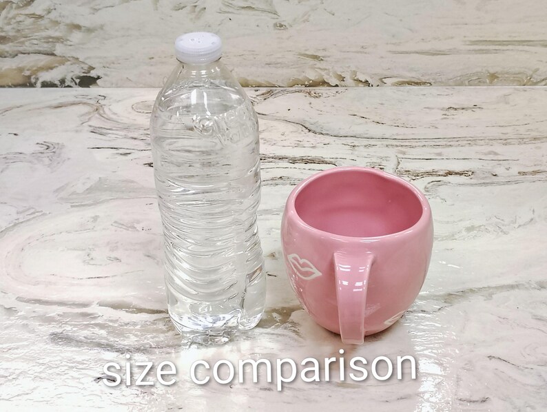 May include: A clear plastic water bottle with a white cap stands next to a pink ceramic mug with a handle. The mug has a white lip design. The text "size comparison" is at the bottom.