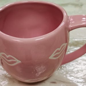 May include: A pink ceramic mug with a curved handle and a wide opening. The mug features two white lip designs on the exterior. The interior of the mug is also pink. The mug is designed for beverages.