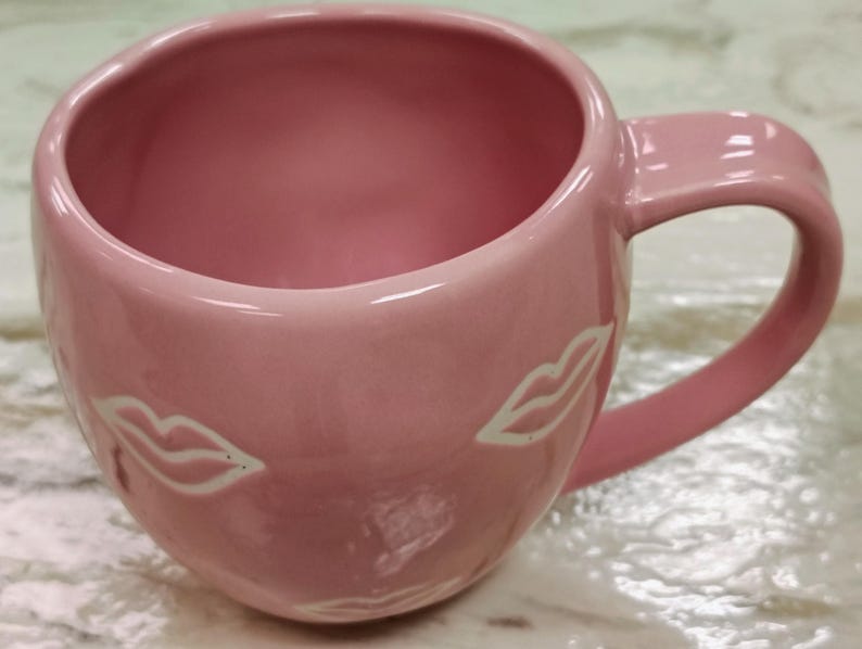 May include: A pink ceramic mug with a curved handle and a wide opening. The mug features two white lip designs on the exterior. The interior of the mug is also pink. The mug is designed for beverages.