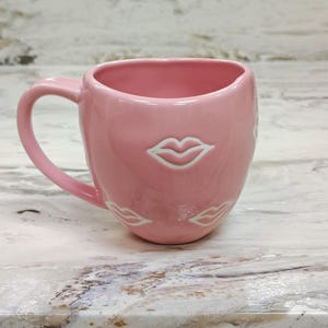 May include: A pink ceramic mug with a curved handle. The mug features a glossy finish and is adorned with white lip designs. The mug is designed with a wide opening and a rounded base, suitable for hot beverages.