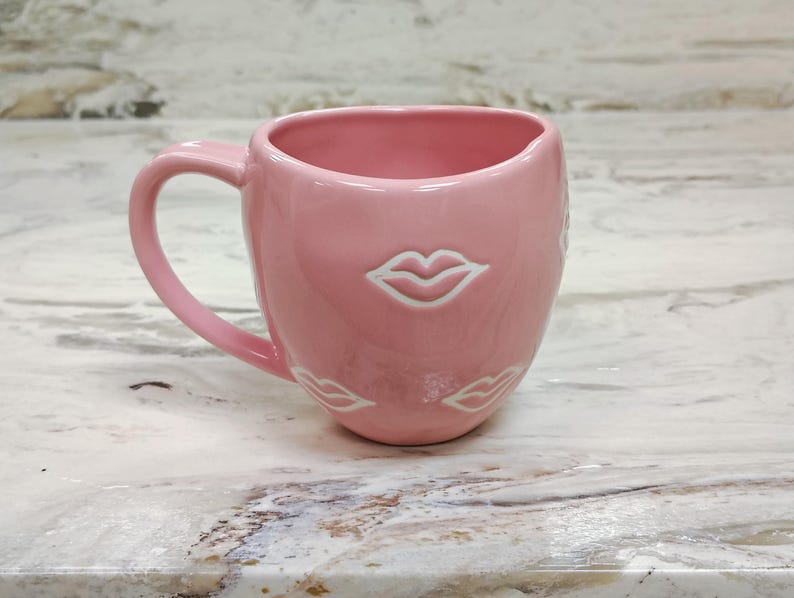 May include: A pink ceramic mug with a curved handle. The mug features a glossy finish and is adorned with white lip designs. The mug is designed with a wide opening and a rounded base, suitable for hot beverages.