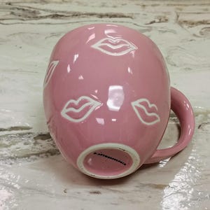 May include: A pink ceramic mug with white lip illustrations. The mug has a rounded shape with a handle and is resting on a marble surface. The bottom of the mug has the text "ANTHROPOLOGIE".