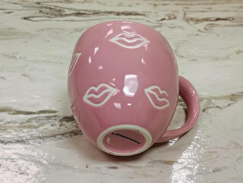 May include: A pink ceramic mug with white lip illustrations. The mug has a rounded shape with a handle and is resting on a marble surface. The bottom of the mug has the text "ANTHROPOLOGIE".