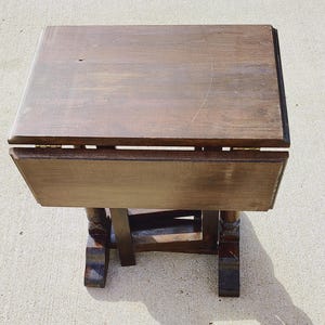 May include: A small, antique wooden drop-leaf table with a dark brown finish. The table has a rectangular top with a hinged leaf on each side. The legs are intricately carved and the table is set on a light-colored surface.
