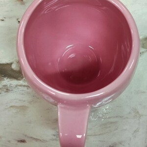 May include: A pink ceramic mug with a rounded body and a single handle. The mug has a glossy finish and is viewed from above, showcasing its interior and handle. The color is a soft, muted pink.