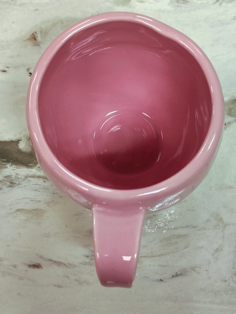 May include: A pink ceramic mug with a rounded body and a single handle. The mug has a glossy finish and is viewed from above, showcasing its interior and handle. The color is a soft, muted pink.