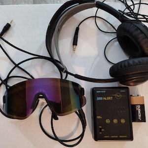 Op de afbeelding: Een verzameling elektronische apparaten: zwarte koptelefoon, spiegelende zonnebril en een zwart apparaat met de tekst "DAVID ALERT". Er is ook een Duracell-batterij aanwezig.