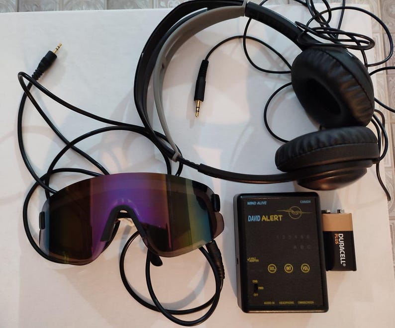 Op de afbeelding: Een verzameling elektronische apparaten: zwarte koptelefoon, spiegelende zonnebril en een zwart apparaat met de tekst "DAVID ALERT". Er is ook een Duracell-batterij aanwezig.