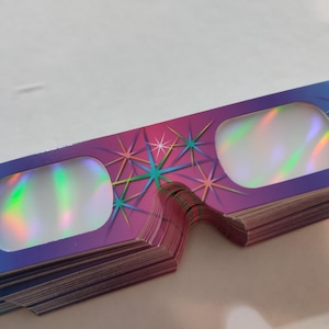 Op de afbeelding: Een stapel 3D-brillen met een kleurrijk ontwerp. De monturen lopen over van blauw naar paars, versierd met sterrenpatronen. De lenzen hebben een holografisch, iriserend uiterlijk.