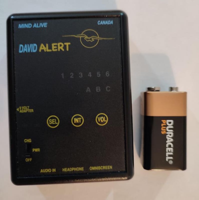 Op de afbeelding: Een zwart "DAVID ALERT"-apparaat met de tekst "MIND ALIVE" en "CANADA". Het heeft knoppen met de labels "SEL", "INT" en "VOL", en een 9-volt adapteringang. Een Duracell Plus 9-volt batterij staat naast het apparaat.