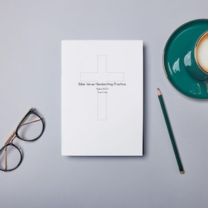 May include: A flat lay image featuring a white notebook with a cross graphic and the text "Bible Verse Handwriting Practice." A teal teacup with gold trim and a pencil are next to the notebook. A pair of glasses is also in the image.