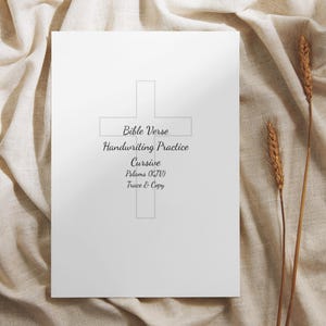 May include: A white sheet of paper featuring a cross outline and the text "Bible Verse Handwriting Practice Cursive Psalms (KJV) Trace & Copy". The paper rests on beige fabric, with wheat stalks.