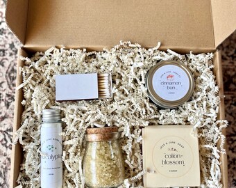 May include: A gift box filled with bath and body products. The box contains a small candle labeled "cinnamon bun", a bottle of bath salts labeled "eucalyptus", a bar of soap labeled "cotton blossom", and a pack of matches.