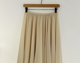 May include: A knee-length skirt in a solid beige color, featuring a lightweight, semi-sheer fabric with a gathered, elasticized waistband. The skirt hangs in soft, vertical pleats from the waist, creating a full, flowing silhouette.