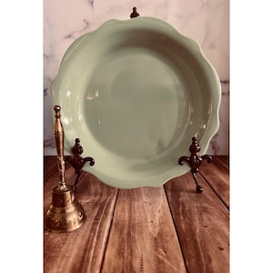 May include: A light sage green ceramic pie dish with a scalloped edge, displayed on a dark metal stand. Beside the dish sits a tall, slender brass hand bell featuring intricate engraved patterns along its flared base and cylindrical handle.
