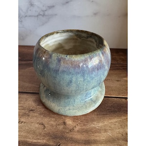 May include: A handmade ceramic bowl with a unique shape and a glazed finish. The bowl features a rounded body with a flared base and a wide opening. The glaze has a blend of blue, green, and brown hues, creating a textured appearance. The bowl sits on a wooden surface.