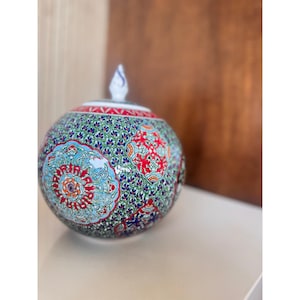 May include: A white ceramic mug with a black handle and the text "But First, Coffee" in a bold, black font. The mug is set against a blurred background, suggesting a cozy setting.