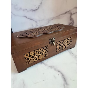 Könnte beinhalten: Eine dunkelbraune Holzbox mit dekorativen Elementen. Der Deckel hat ein lasergeschnittenes Design, und die Vorderseite hat zwei Abschnitte mit kreisförmigen Holzakzenten. Ein Metallverschluss sichert die Box, geeignet für Aufbewahrung oder Präsentation.