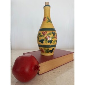 Può includere: Una bottiglia in ceramica gialla con tappo in sughero, decorata con foglie verdi e bacche rosse dipinte a mano in fasce orizzontali. La bottiglia poggia su un libro rosso spesso e rettangolare accanto a un frutto di melograno rosso liscio.