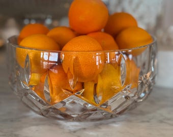 May include: A clear, cut-glass bowl filled with bright orange oranges. The bowl has a diamond pattern and sits on a marble surface. The oranges are ripe and have a textured peel.