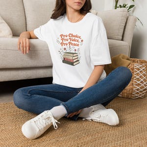 以下が含まれることがあります： 白い半袖Tシャツ。胸の中央に、花柄のアクセントが付いた4冊の本の積み重ねのグラフィックと、「Pro Choice, Pro Voice, Pro Peace」という錆色のテキストがプリントされています。