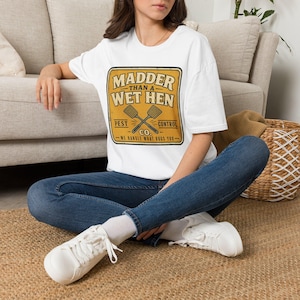 Può includere: Una t-shirt bianca a maniche corte con una grafica quadrata gialla che riporta il testo MADDER THAN A WET HEN PEST CONTROL CO. WE HANDLE WHAT BUGS YOU e un'illustrazione di due scacciamosche incrociati al centro del design.