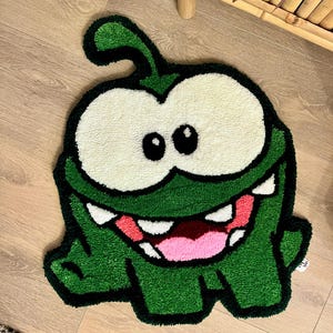 Pode incluir: Um tapete verde em estilo cartoon em forma de personagem com olhos grandes brancos, boca rosa e dentes brancos. O tapete tem um corpo verde com contorno preto. O tapete é colocado em um piso de madeira.
