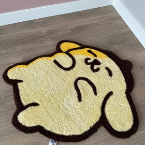 May include: A cartoon-shaped rug with a yellow head and body, brown outline, and black facial features. The rug features the text "tuff boi" in the bottom left corner. The rug is designed for floor use.