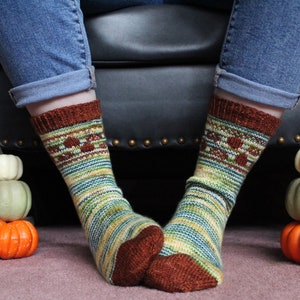May include: A pair of colorful knitted socks with a brown, green, and yellow pattern. The socks are worn by a person sitting on a black leather chair. There are three small pumpkins stacked on either side of the chair.