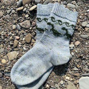 May include: A pair of gray knitted socks with a green wave pattern. The socks are lying on a bed of small gray and brown stones.