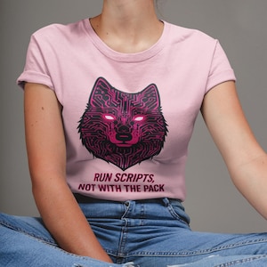 Puede incluir: Una camiseta rosa claro con un diseño de lobo negro y rosa, con ojos brillantes, construido con patrones de circuitos. El texto "RUN SCRIPTS, NOT WITH THE PACK" está impreso debajo. La camiseta se combina con unos vaqueros azules.