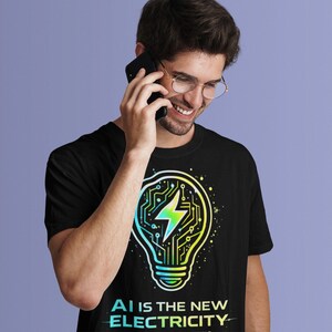 May include: Black t-shirt with a lightbulb design incorporating a circuit board pattern and a lightning bolt in green, yellow, and blue. The text "AI IS THE NEW ELECTRICITY" is printed below the graphic.