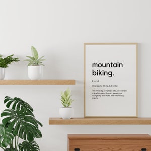 以下が含まれることがあります： モダンなフォントで「mountain biking」という言葉が書かれた額入りのプリント。マウンテンバイクの定義がタイトルの下にあります。プリントは、鉢植えの植物が置かれた木製の棚に飾られています。