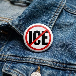 May include: A round white button with the words "ICE" in black, crossed out with a red line, resting on a blue denim jacket. The button has a red border. The denim jacket has visible stitching and a button closure.