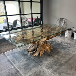 May include: A dining table featuring a large, natural light brown tree root base with an organic, gnarled texture. A rectangular, clear glass top rests directly on the wooden root structure, providing a flat surface for use.