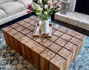May include: A large wooden coffee table made of stacked square blocks. The table is in a living room setting with a white couch and a vase of pink and white flowers on top.