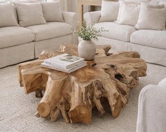 May include: A coffee table crafted from a large, natural light brown tree root with a smooth, flat top surface. A light-colored ceramic vase containing green foliage and two stacked books are placed on the center of the wooden root table.