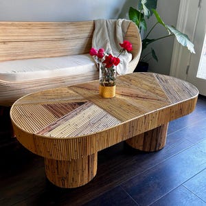 May include: A woven rattan coffee table with a unique geometric design. The table has two round legs and a rectangular top. A vase with red flowers sits on the table.