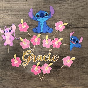May include: A collection of colorful party decorations featuring the characters Stitch and Angel, along with pink hibiscus flowers and the name "Gracie" in gold script. The decorations are arranged on a wooden surface.