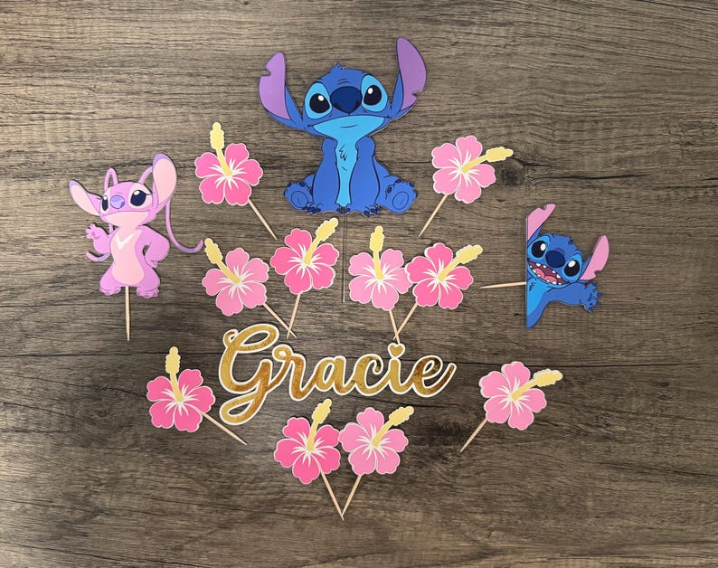 May include: A collection of colorful party decorations featuring the characters Stitch and Angel, along with pink hibiscus flowers and the name "Gracie" in gold script. The decorations are arranged on a wooden surface.