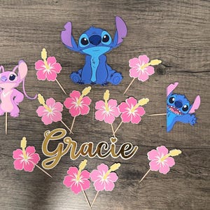 May include: A collection of themed cake toppers featuring the characters Stitch and Angel, along with pink hibiscus flowers and the name "Gracie" in gold script. The toppers are made of paper and attached to wooden sticks.