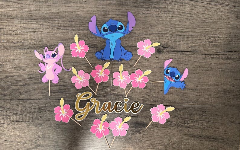 May include: A collection of themed cake toppers featuring the characters Stitch and Angel, along with pink hibiscus flowers and the name "Gracie" in gold script. The toppers are made of paper and attached to wooden sticks.
